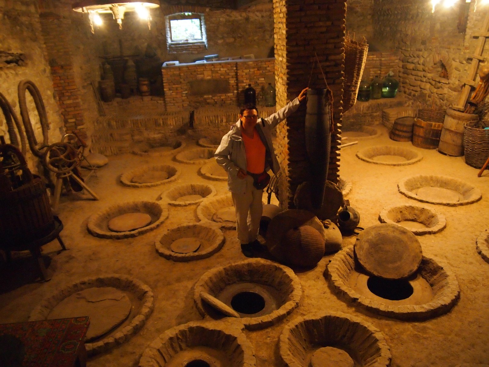 A traditional Georgian marani (wine cellar) in Kakheti, with dozens of qvevri clay wine vessels buried in the earthen floor, their round lids visible at floor level. A person stands at the center for scale.